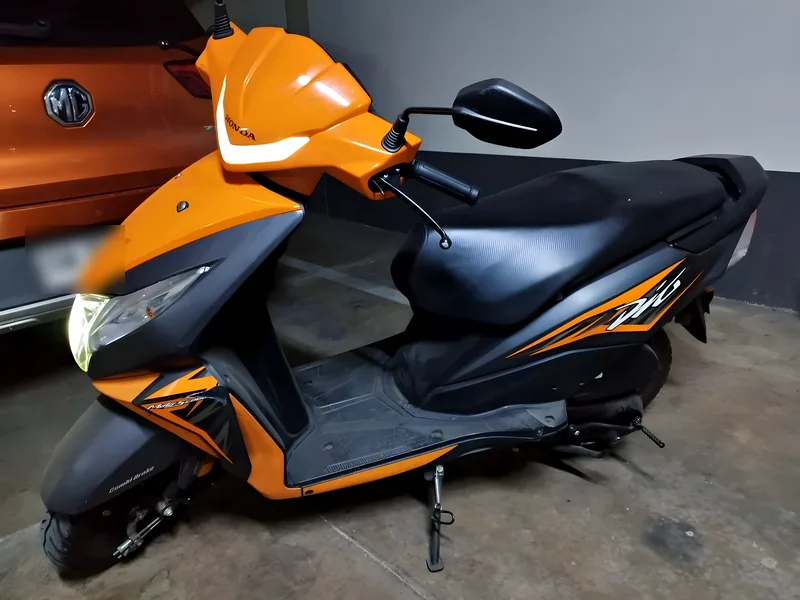Foto real de la Honda Dio que se rifa, color naranjo con negro, estacionada.